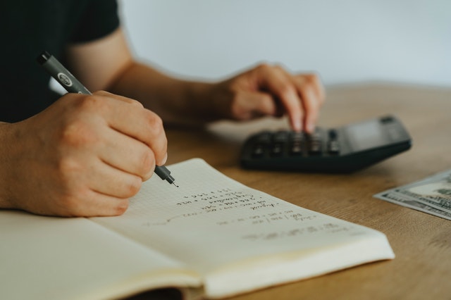 Close up of a person sitting at a desk, using a calculator with their left hand and holding a pen while writing in a notebook with their right hand.